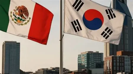 Mexican and South Korean flags waving above a city skyline with tall buildings behind them.