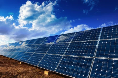 Solar panels angled on metal frames in a dry grassy field beneath a bright blue sky with clouds