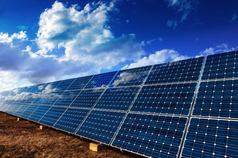 Solar panels angled on metal frames in a dry grassy field beneath a bright blue sky with clouds