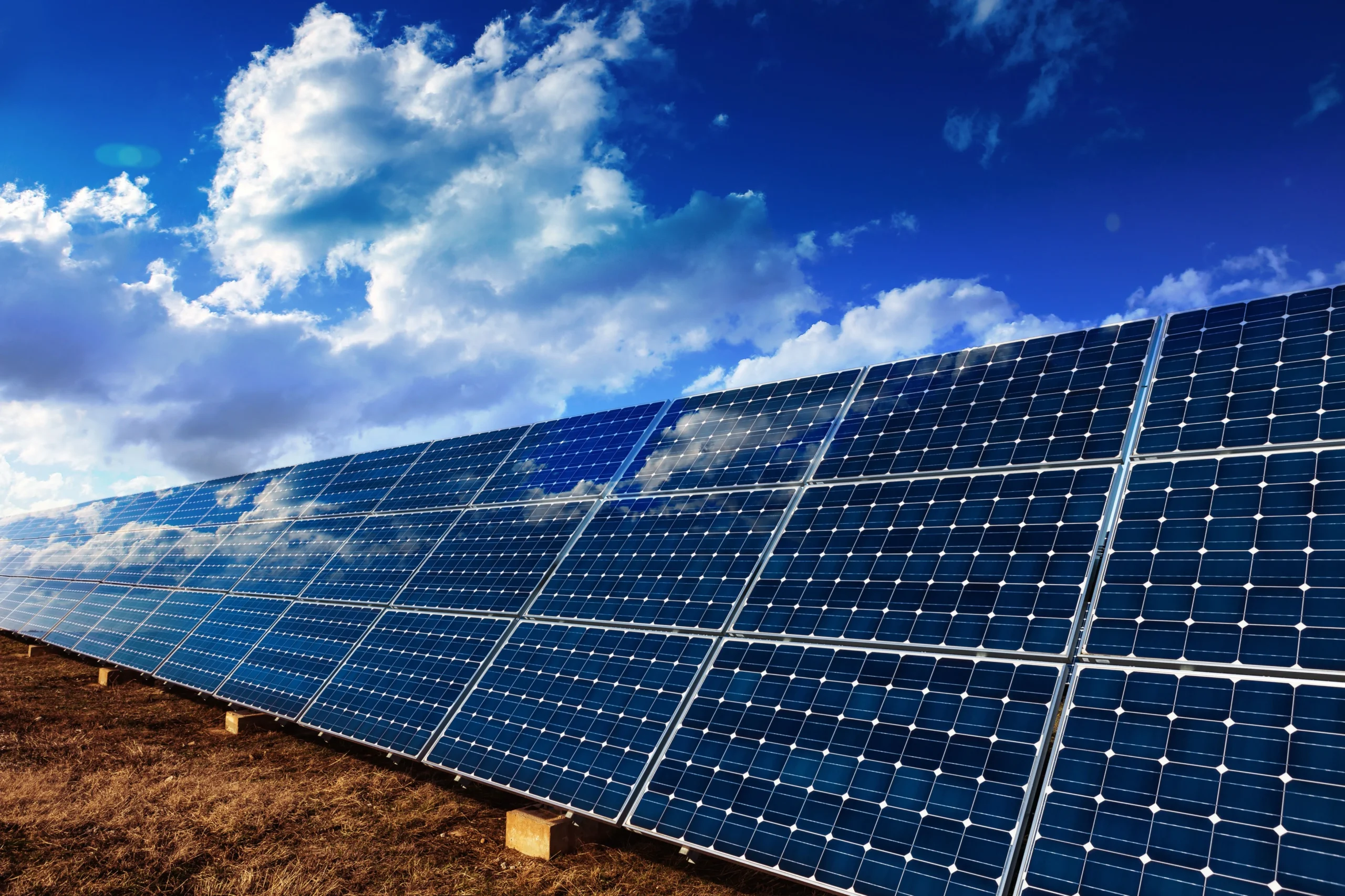 Solar panels angled on metal frames in a dry grassy field beneath a bright blue sky with clouds