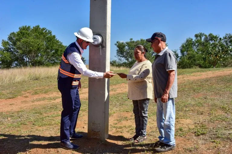 Worker in a white hard hat and safety vest hands a document to two adults beside a concrete utility pole in an open field.