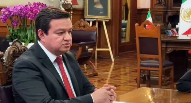 Man in a dark suit and red tie sits at a conference table, hands folded, in a formal office with a Mexican flag in the background.