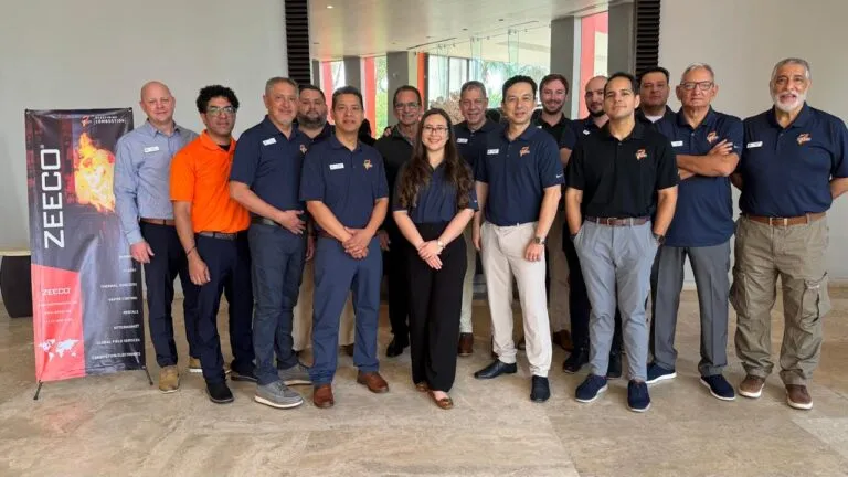 Group of professionals in navy polo shirts (one in orange) posing for a company photo indoors beside a Zeeco banner, smiling at the camera.