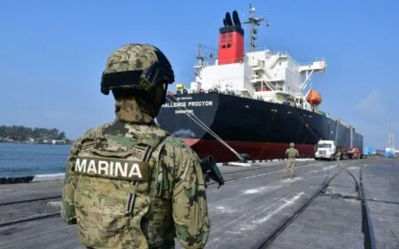Soldier in camo with a ‘MARINA’ patch standing on a port quay, facing a large black and red cargo ship.