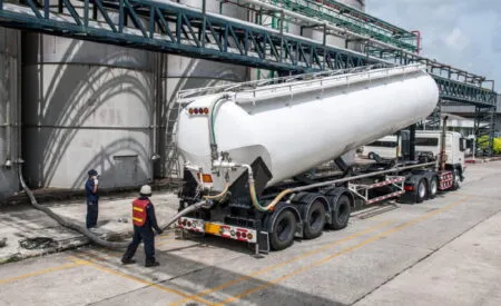 White tanker trailer connected to hoses at an industrial facility, with workers in safety gear nearby under metal framework and storage tanks nearby