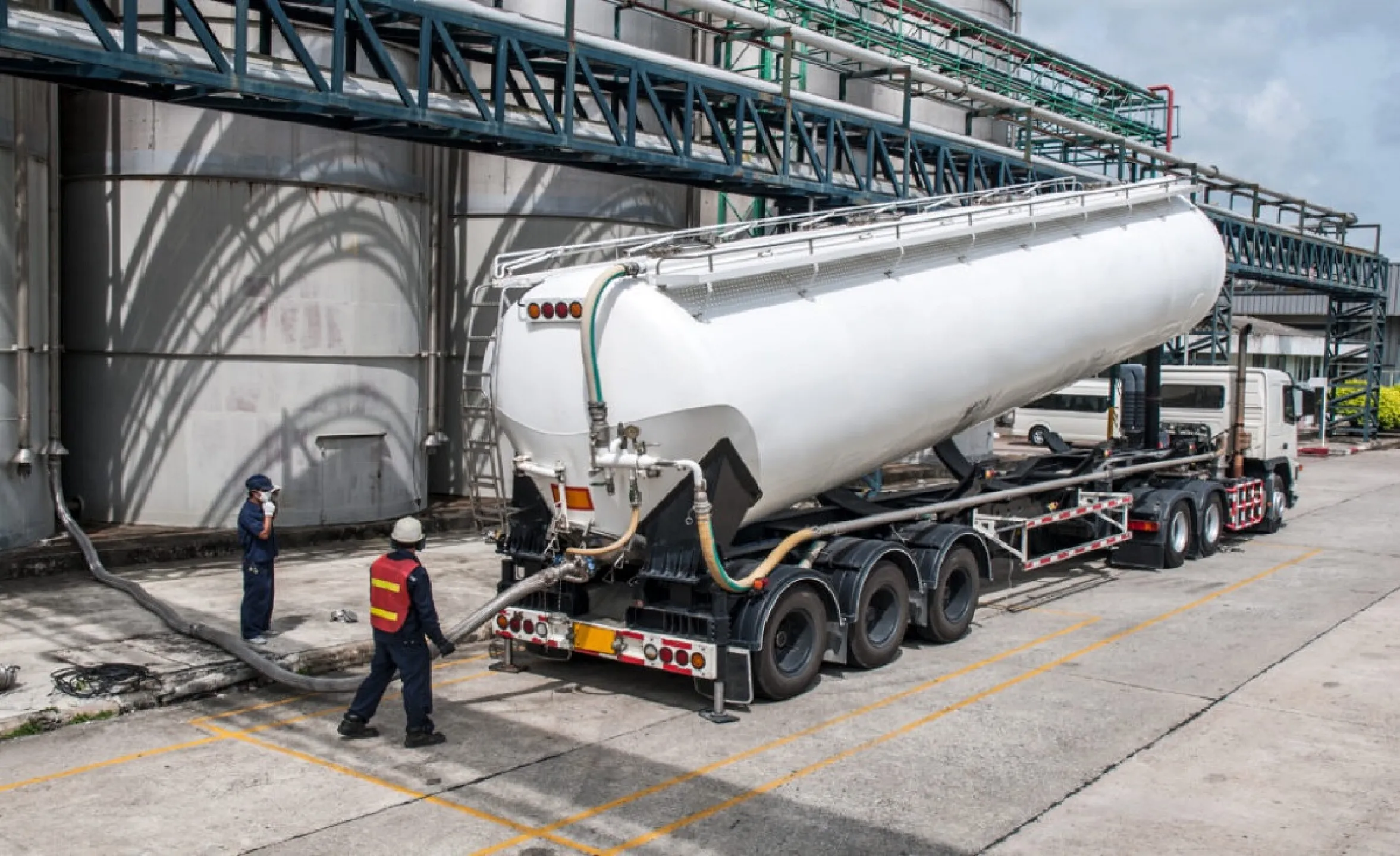 White tanker trailer connected to hoses at an industrial facility, with workers in safety gear nearby under metal framework and storage tanks nearby