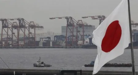 Japanese flag waving in the foreground as container port cranes rise over the harbor with ships in the background.