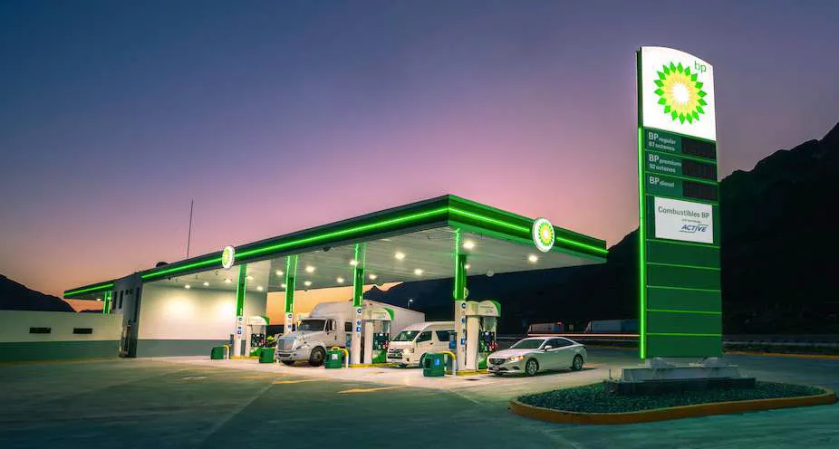 BP gas station at dusk with multiple fueling pumps and a tall illuminated sign, two trucks and a car refueling under a green-lit canopy