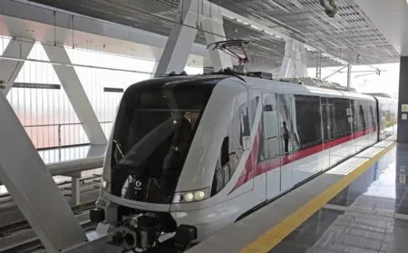 Sleek white light-rail train at a modern station with a yellow tactile edge on the platform and overhead catenary equipment above the vehicle.