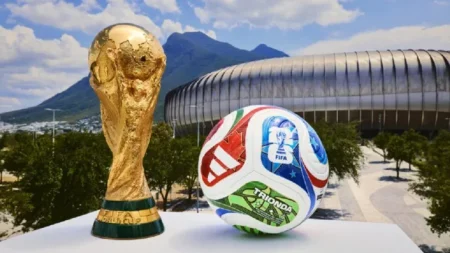 Gold FIFA World Cup trophy beside a colorful football on a pedestal, with a stadium and mountains in the background.