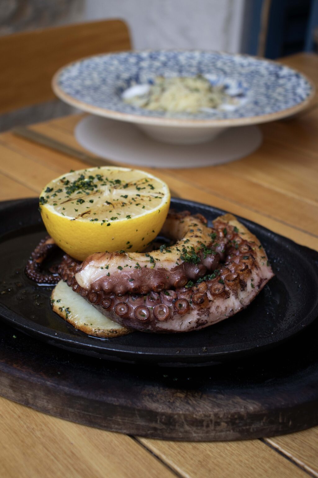 00590 - Petróleo y Energía Grilled octopus tentacle on a dark plate with a halved lemon, sprinkled with herbs, on a wooden table.