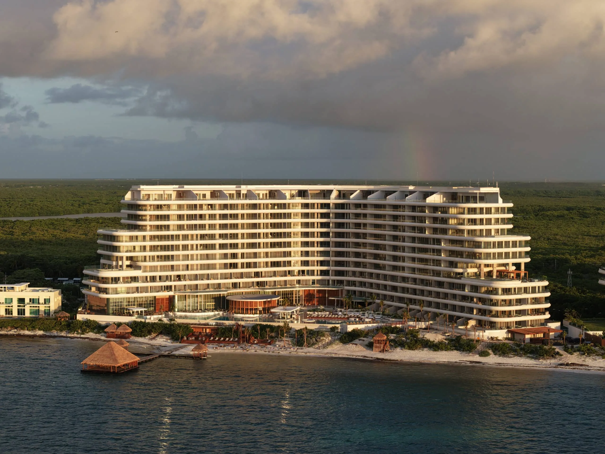 Modern white beachfront hotel with curved balconies, ocean in foreground and a rainbow in the cloudy sky.