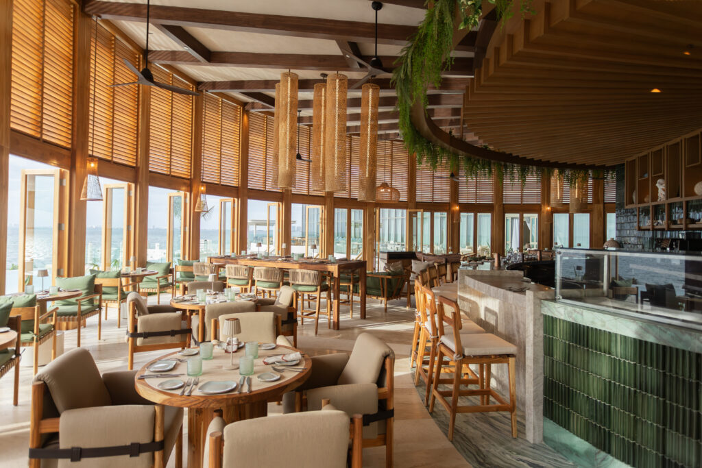 04 - Petróleo y Energía Sunlit seaside restaurant interior with wooden beams, hanging wicker lights, and green-cushioned seating by large windows overlooking the water.