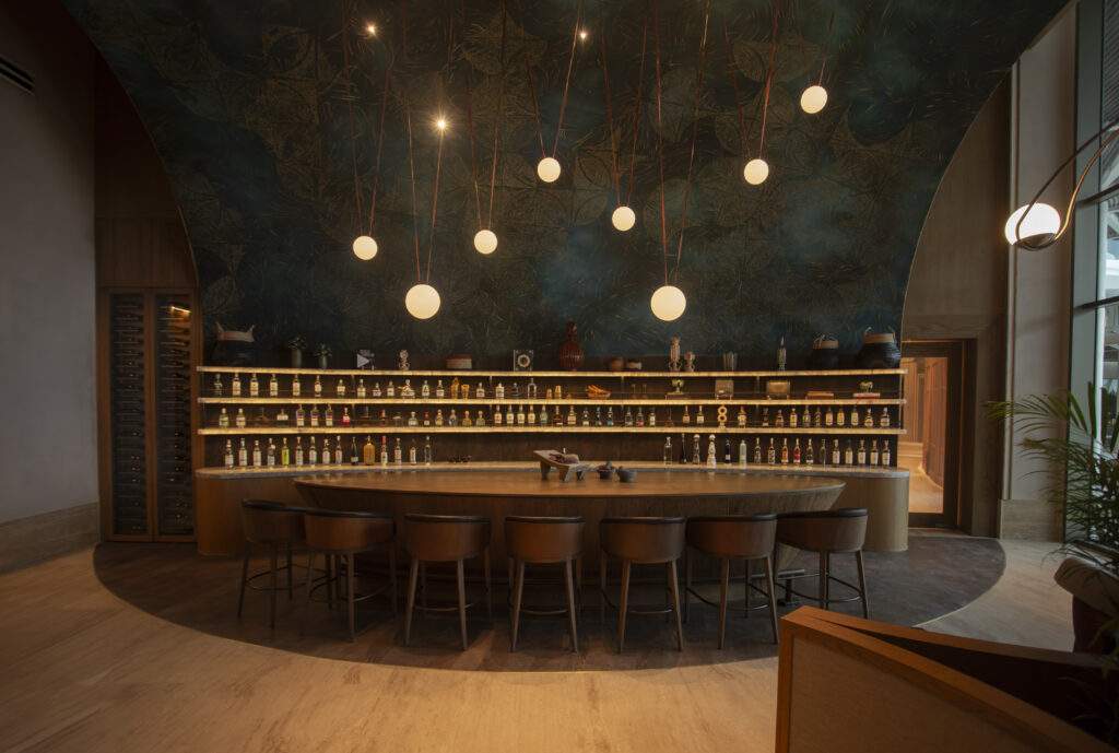 05 - Petróleo y Energía Warm-lit hotel or restaurant bar with a curved wooden counter and shelves filled with bottles against a dark leafy-patterned wall, illuminated by hanging globe lights.