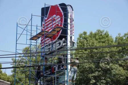 Workers on scaffolding install a tall storefront sign with a red graphic, outdoors under a blue sky, power lines crossing in front of them.