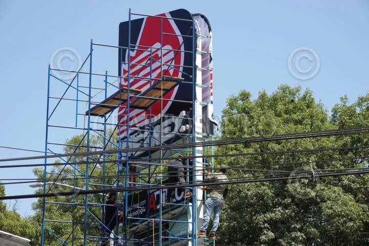 Workers on scaffolding install a tall storefront sign with a red graphic, outdoors under a blue sky, power lines crossing in front of them.