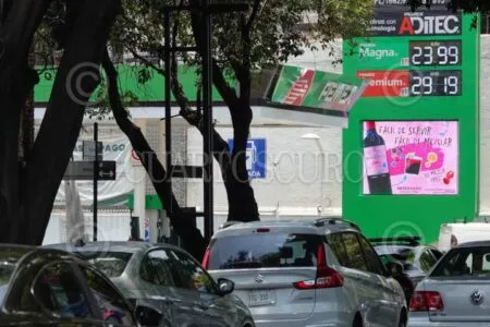 Street view with a bright green gas station storefront, price board showing 23.99 and 29.19, and several cars in traffic amid trees and posters.