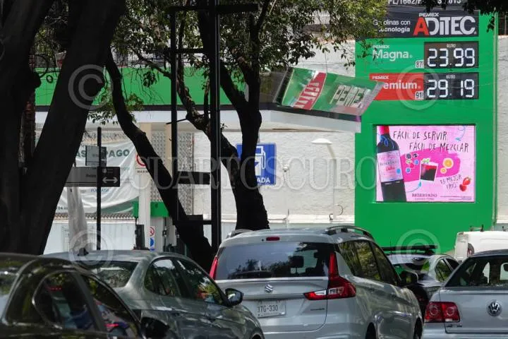 Street view with a bright green gas station storefront, price board showing 23.99 and 29.19, and several cars in traffic amid trees and posters.