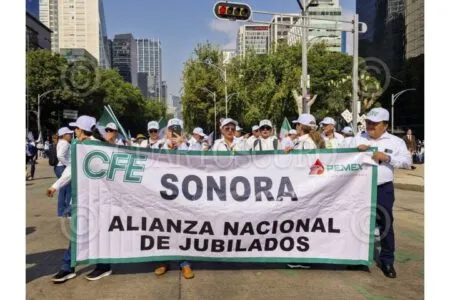 Group of retirees in white hats marching down a city street, holding a banner reading 'CFE SONORA ALIANZA NACIONAL DE JUBILADOS'.