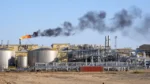 Industrial refinery with large storage tanks, extensive piping, and a flaming flare emitting black smoke.
