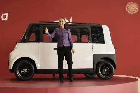 Woman in a purple blazer giving a thumbs-up beside a white compact van on a red exhibition platform.