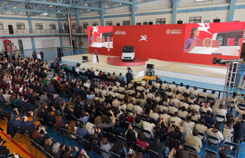 1780926115218 - Petróleo y Energía Large indoor ceremony with a red stage, a small white vehicle on a circular platform, and a seated audience watching the unveiling.