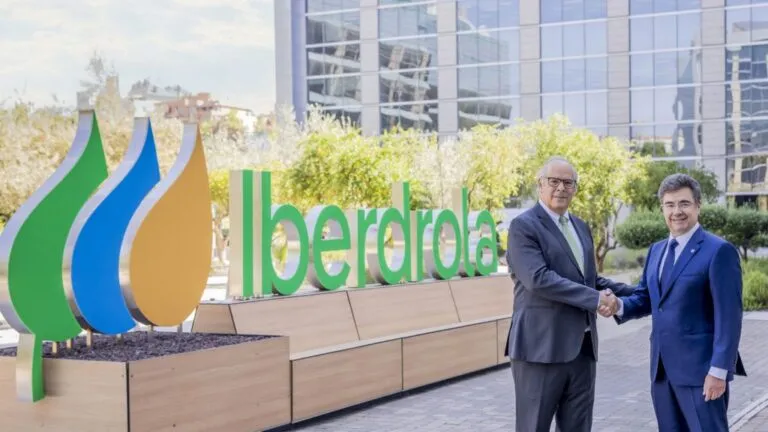 Two men in suits shake hands in front of a large Iberdrola sign at an outdoor corporate plaza.