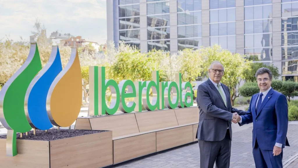 Two men in suits shake hands in front of a large Iberdrola sign at an outdoor corporate plaza.