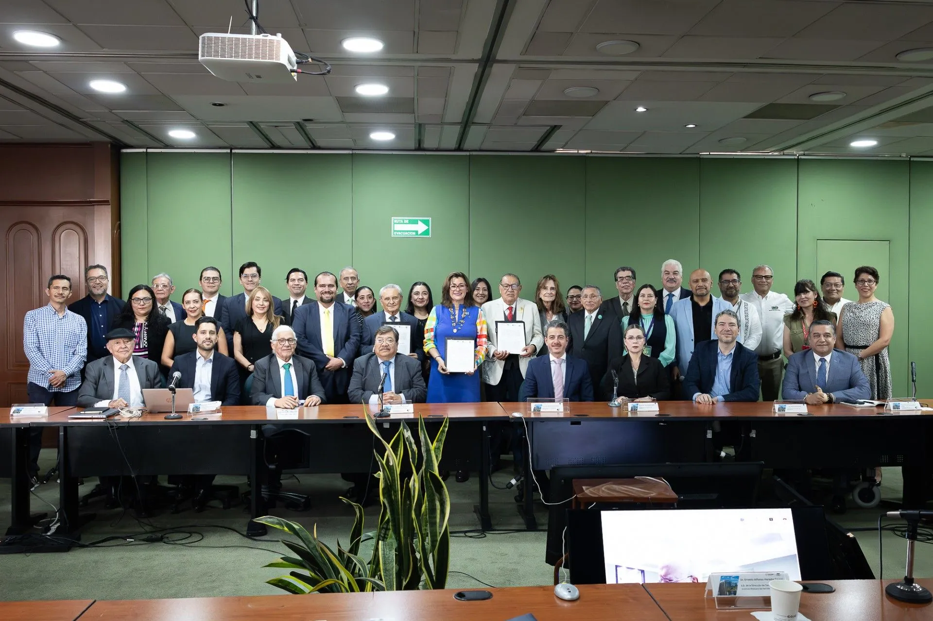 Large group of professionals in a conference room posing for a group photo, with two people in front holding certificates.