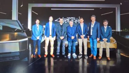Group of seven men in blue blazers posing for a photo in a modern showroom with futuristic cars in the background, stage lighting overhead.
