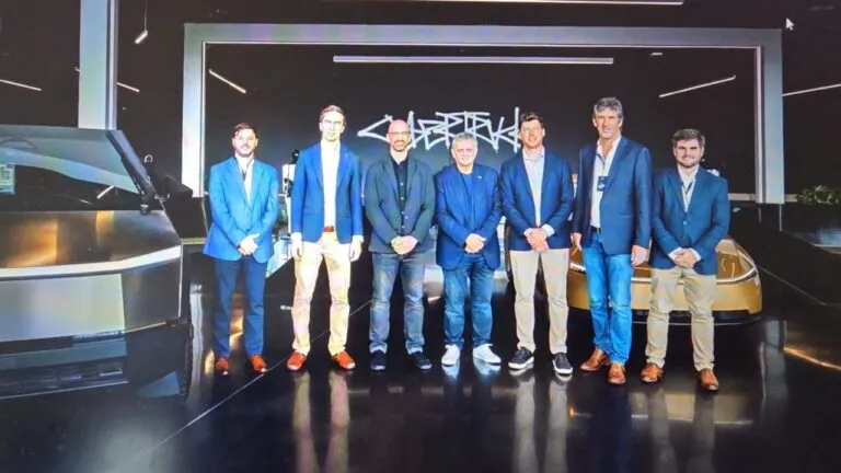 Group of seven men in blue blazers posing for a photo in a modern showroom with futuristic cars in the background, stage lighting overhead.