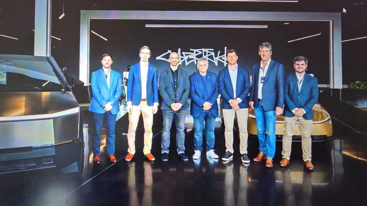 Group of seven men in blue blazers posing for a photo in a modern showroom with futuristic cars in the background, stage lighting overhead.