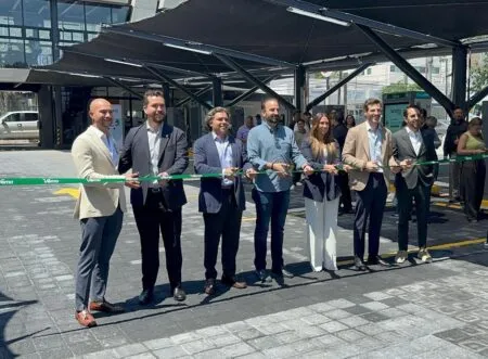 Eight professionally dressed people hold a green ribbon for a ribbon-cutting ceremony outside a glass-front building.