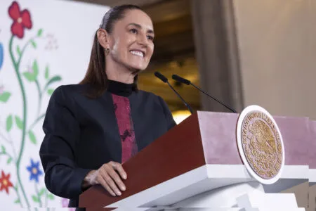 Smiling woman at a lectern with two microphones; an official seal on the front and a floral backdrop behind.