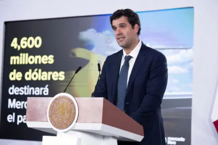 Man in a dark suit speaks at a podium with the Mexican seal; screen behind shows text about 4,600 millones de dólares in Spanish.