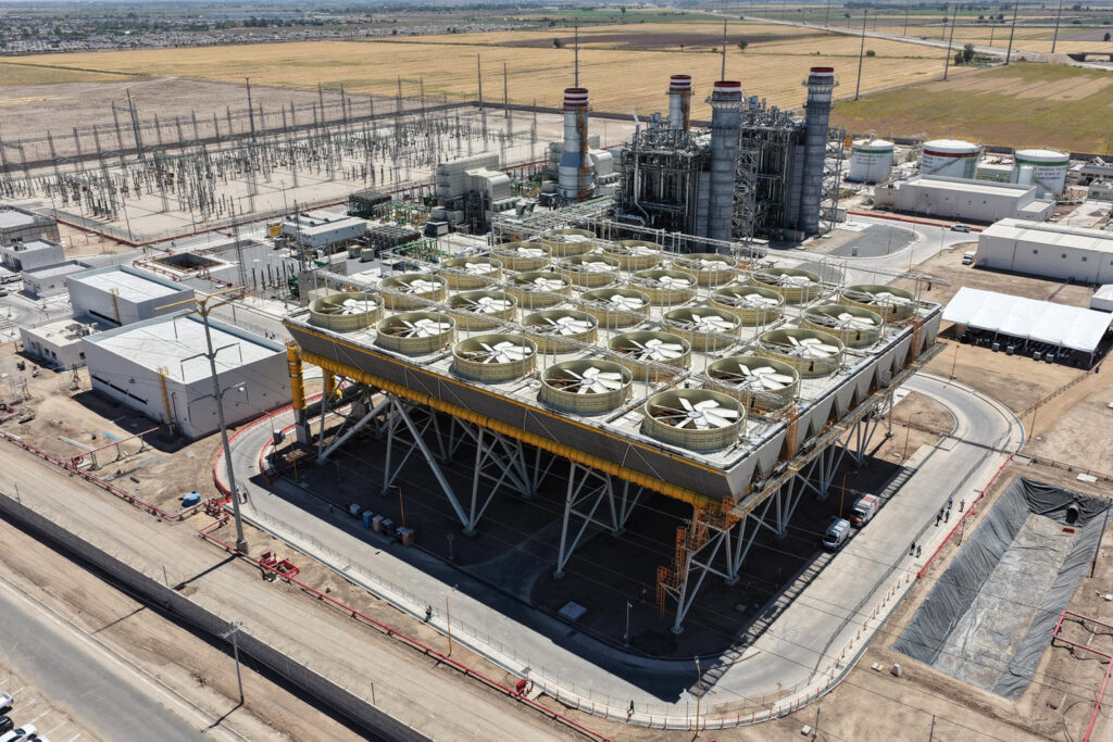 2026-06-21_Inauguraci_n_Central_de_Ciclo_Combinado_Mexicali_28_JLC - Petróleo y Energía Large industrial power plant with a row of cooling fans on a raised platform, surrounded by piping, towers, and electrical infrastructure in a flat desert landscape.