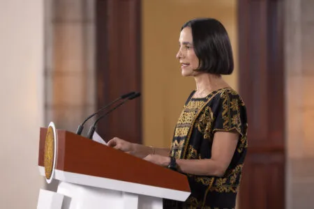 Woman delivering a speech at a podium with two microphones, wearing a black and gold patterned top and looking to the side in a formal setting.