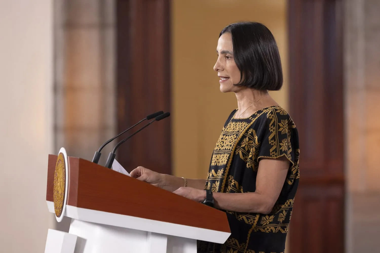 Woman delivering a speech at a podium with two microphones, wearing a black and gold patterned top and looking to the side in a formal setting.