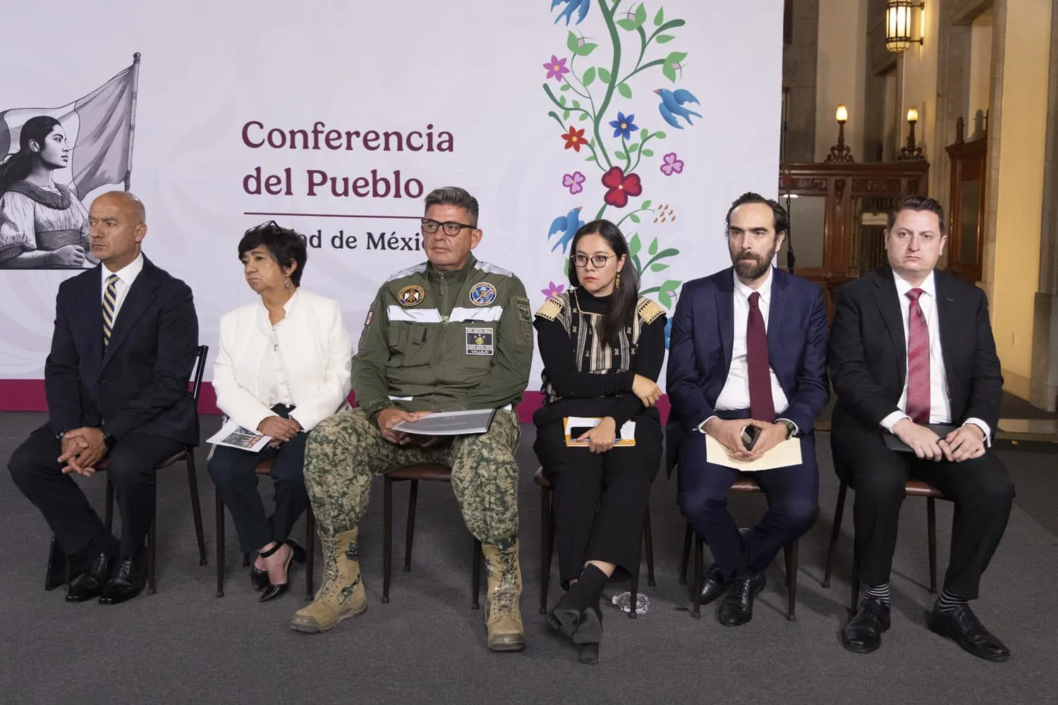 Six officials seated in a row at a conference panel, with a backdrop reading 'Conferencia del Pueblo' and a floral design behind them.