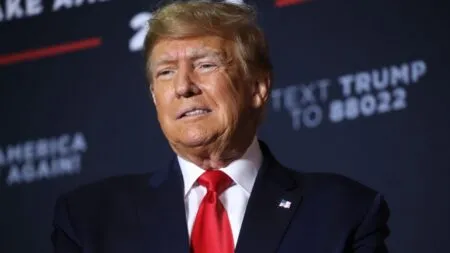 Man in a dark suit with a red tie and an American flag lapel pin, standing on a dark stage backdrop at a political event.