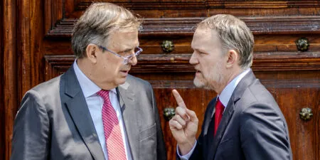 Two suited men conversing closely in front of a wooden door, one raising a finger as if making a point.