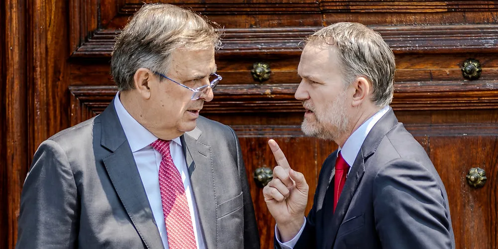 Two suited men conversing closely in front of a wooden door, one raising a finger as if making a point.