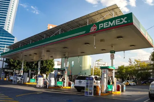 Pemex gas station with a large canopy and multiple pumps; a white SUV refueling under a blue sky in urban surroundings.