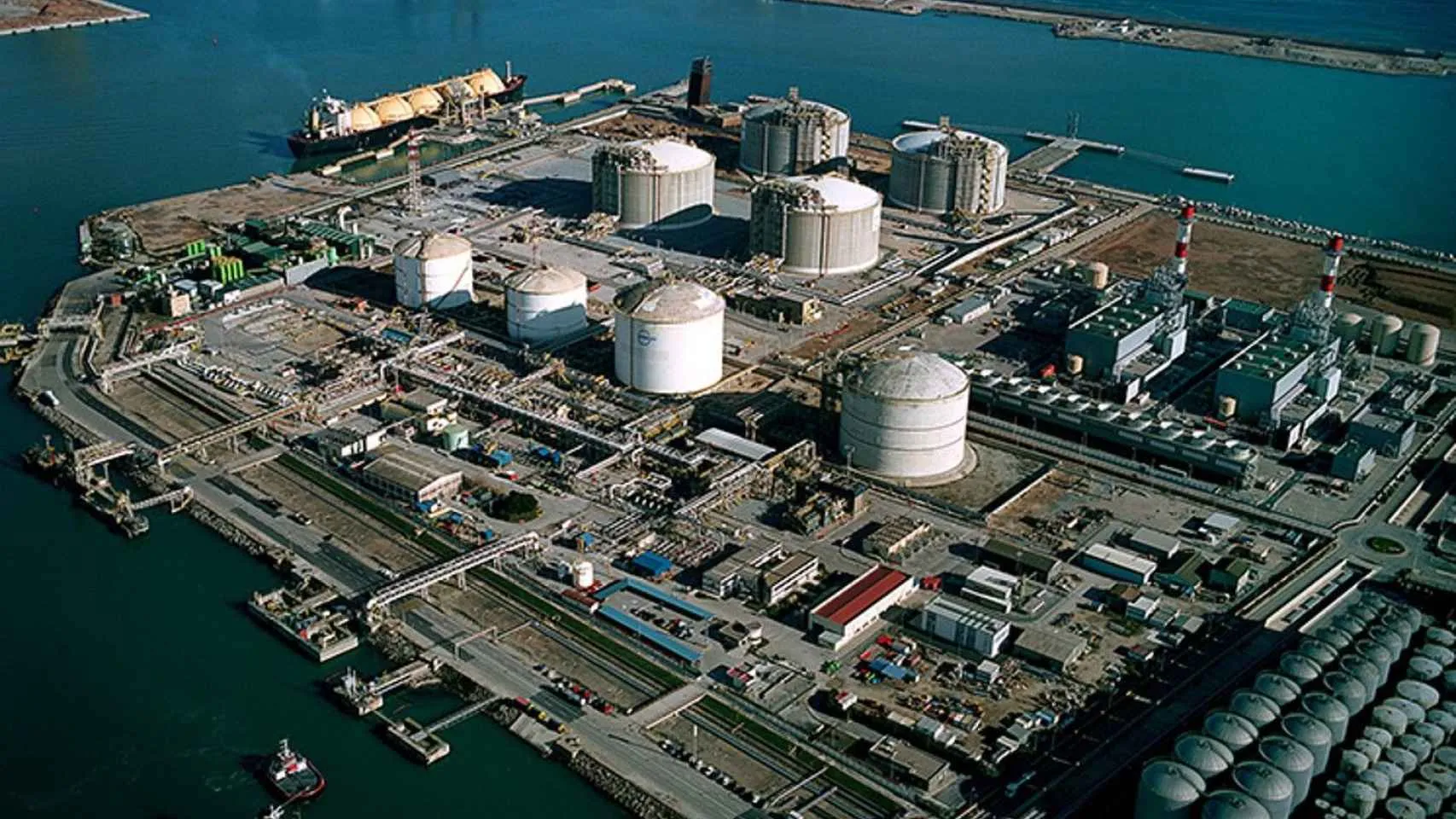 Aerial view of a large coastal industrial complex with numerous storage tanks, pipelines, and support buildings by the harbor