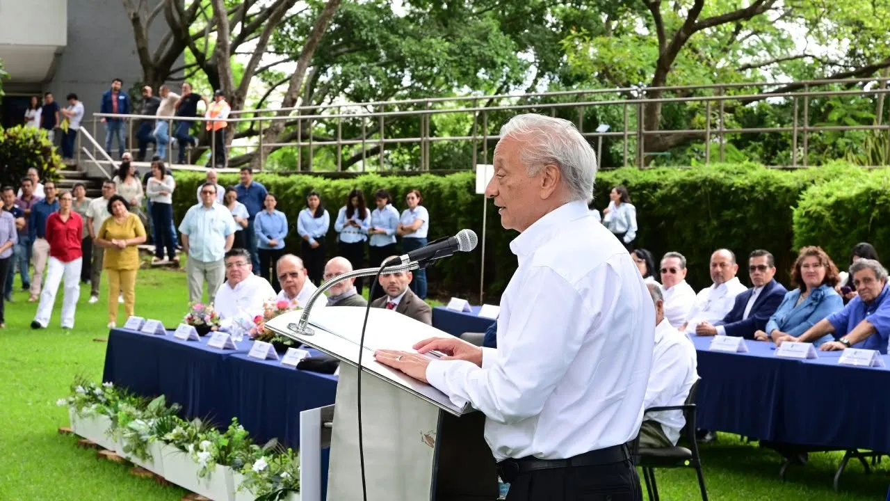Elderly man in white shirt speaks at a podium with a microphone during an outdoor ceremony; a long table of officials sits behind him on a lawn with greenery and attendees nearby.