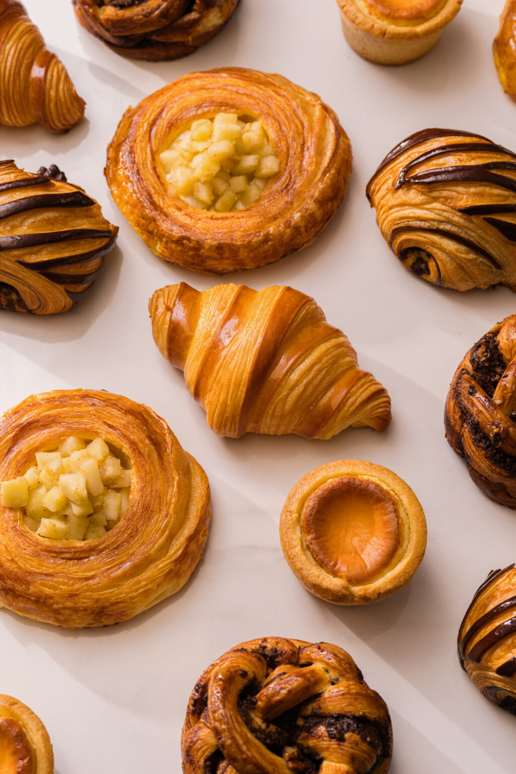 AMEX CL1236 1 - Petróleo y Energía Assorted pastries on a light background: apple Danish, croissants, chocolate-swirl palmiers, and small fruit tarts.