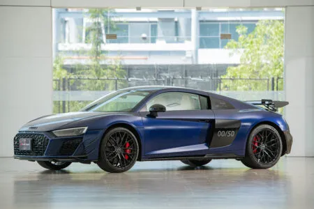 Blue Audi R8 sport coupe in a bright showroom, visible front grille, black wheels, and red brake calipers, with 00/50 marking on the rear door area.