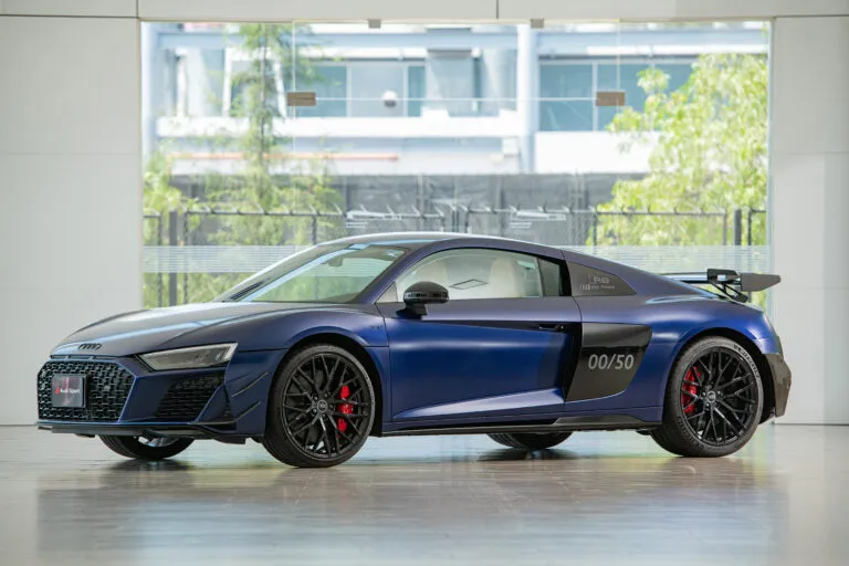 Blue Audi R8 sport coupe in a bright showroom, visible front grille, black wheels, and red brake calipers, with 00/50 marking on the rear door area.