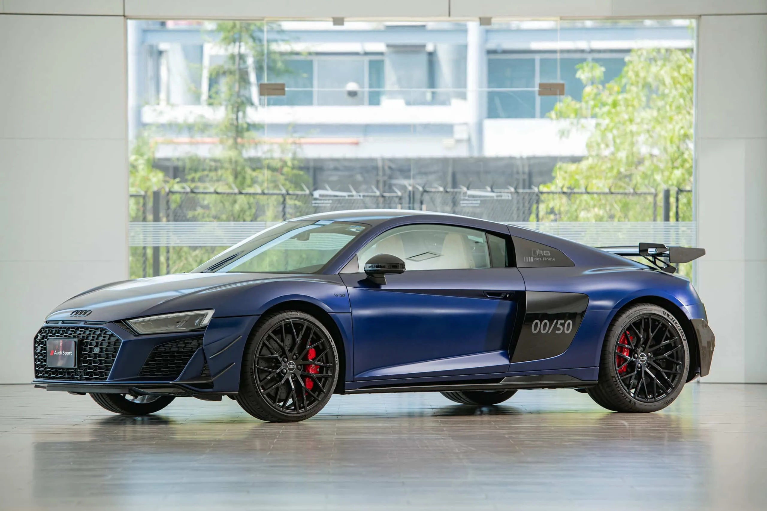Blue Audi R8 sport coupe in a bright showroom, visible front grille, black wheels, and red brake calipers, with 00/50 marking on the rear door area.