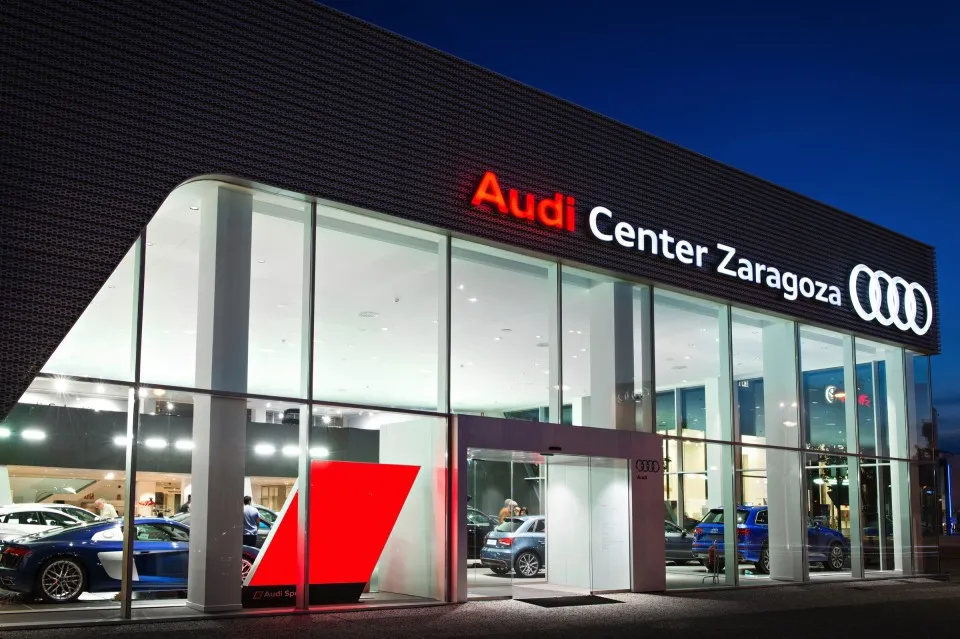 Night view of Audi Center Zaragoza showroom with glass walls, bright interior, and several cars on display.